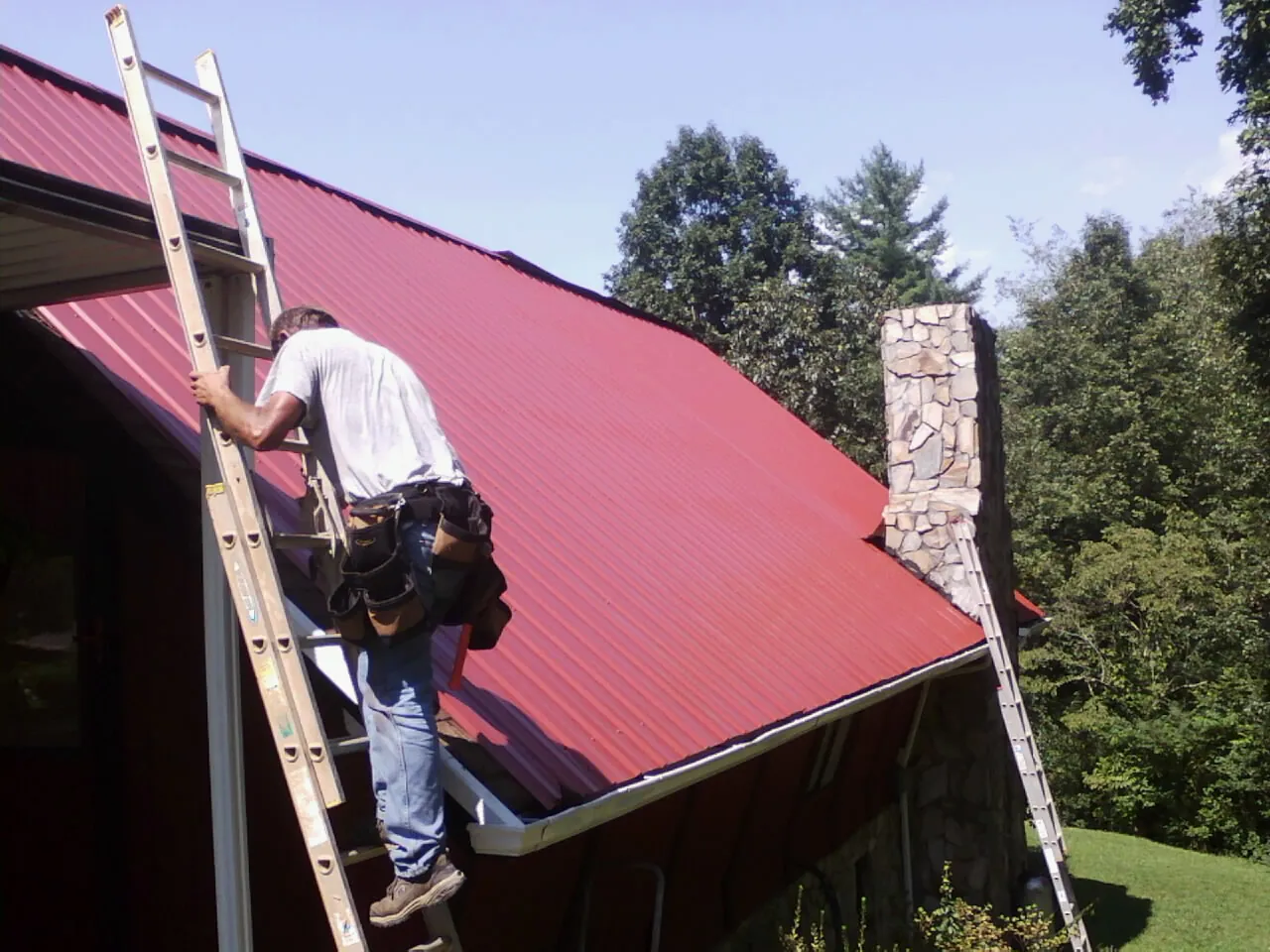 Roofer on a ladder beside a red metal roof and stone chimney — Soffit & Fascia Repair by Roofing Brothers in Speer